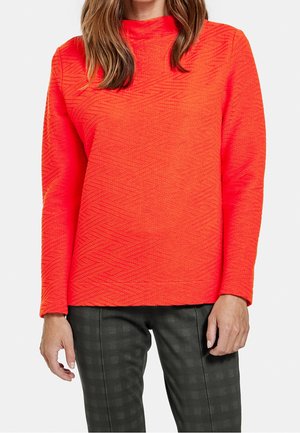 Woman wearing a bright orange textured long-sleeve top and dark checkered pants from shoulders to hips against a plain background.