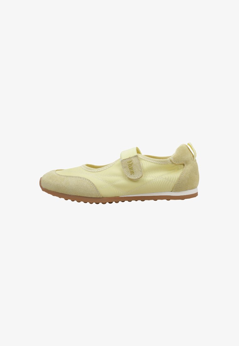 Zapato plano casual amarillo claro con tira de velcro, parches de ante en la punta y el talón, y suela de goma con pequeñas ranuras.