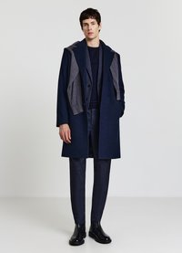 Cappotto di lana blu navy con dettaglio cappuccio grigio, chiusura con bottoni e due tasche laterali. Indossato sopra un completo testurizzato blu navy e stivaletti neri alla caviglia.