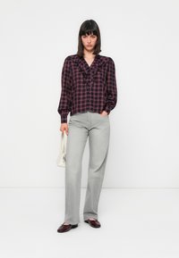 Plaid shirt with ruffles in dark red and navy, paired with light gray wide-leg pants. Model holds a white handbag and wears burgundy shoes.