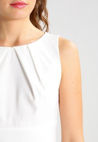 Weißes ärmelloses Kleid mit eingekräuseltem Ausschnitt, glatter Textur und tailliertem Oberteil. Das Design ist minimalistisch und elegant.