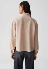 Light beige long-sleeve blouse with a textured fabric, slightly relaxed fit, standing collar, and elastic cuffs, paired with dark jeans.