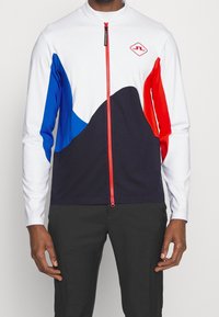 Långärmad zip-up topp med vit kropp, blå och röda detaljer samt marinblå nedre del. Har en hög krage och röd dragkedja.