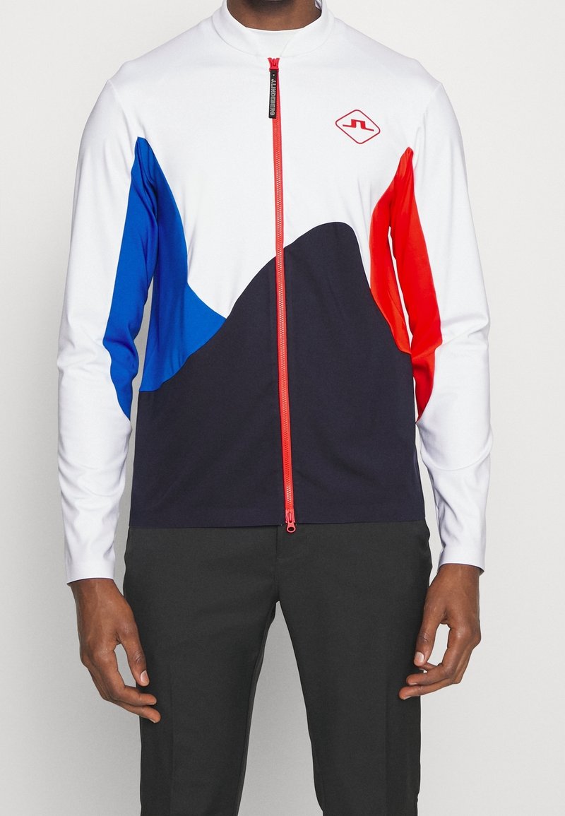 Långärmad zip-up topp med vit kropp, blå och röda detaljer samt marinblå nedre del. Har en hög krage och röd dragkedja.