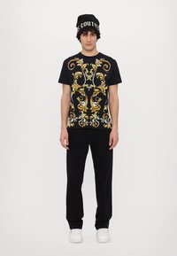 Camiseta preta com um intrincado padrão floral amarelo e branco, combinada com calças pretas e ténis brancos, usada com um boné preto.