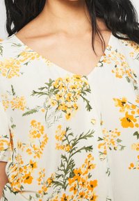 Blouse fleurie avec un décolleté en v, des manches courtes et des motifs floraux jaunes sur un fond clair avec des accents verts et des boutons.