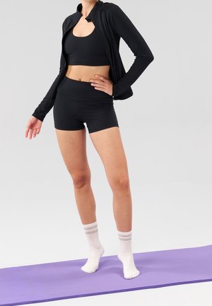 Woman wearing black sportswear and white socks stands on a purple yoga mat with one hand on her hip against a plain background.