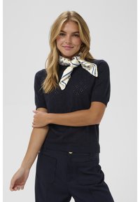 Šviesiaplaukė moteris dėvi tamsiai mėlyną trumpomis rankovėmis megztą palaidinę ir kelnes, aplink kaklą surištą baltai mėlyną šaliką su raštu.