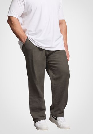 Mand iført løstsiddende mørkegrå bukser, hvid t-shirt, hvide sneakers, hænderne i lommerne, stående mod en ensartet lys baggrund.