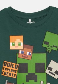 T-shirt en coton vert avec des personnages en pixel art aux formes carrées, incluant un creeper vert et du texte disant "CONSTRUIRE EXPLORER CRÉER."