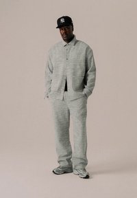 Casaco texturado cinzento e calças correspondentes, com um corte descontraído, fecho de botões visível, combinados com ténis cinzentos e um boné preto.