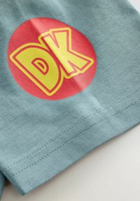 T-shirt en coton bleu clair avec un graphique rond rouge portant les lettres "DK" en jaune, en police gras. Couture visible à l'ourlet.
