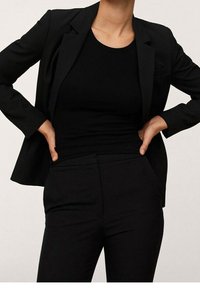 Blazer noir sur mesure porté sur un débardeur noir côtelé, associé à un pantalon noir ajusté. L'ensemble présente un design élégant et sans couture.