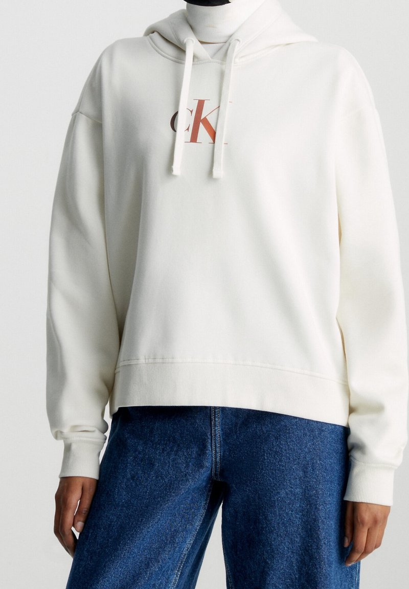 Person wearing a white hoodie with drawstrings and a small "CK" logo, paired with blue denim jeans, against a plain background.