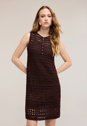 Jonge vrouw met lang golvend haar, gekleed in een mouwloze, donkerbruine gehaakte jurk met knoopdetails op een neutrale achtergrond.