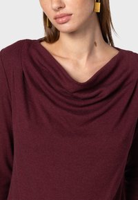 Bordowy top z dekoltem w łódkę, wykonany z miękkiego materiału, z podwieszanym przodem i długimi rękawami, o gładkiej teksturze i dopasowanym fasonie.