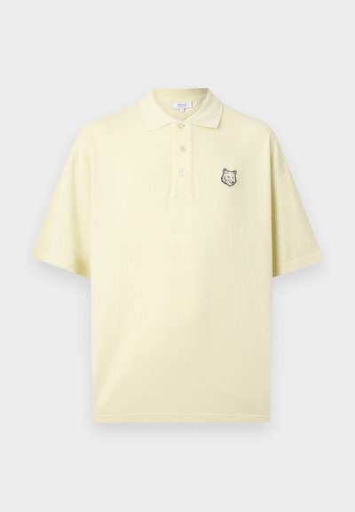 Απαλό κίτρινο κοντομάνικο πόλο με γιακά, τρία κουμπιά και μικρό μαύρο λογότυπο κεφαλής λύκου στο αριστερό στήθος.