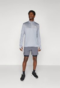 Lichtgrijs sportshirt met een kwart-ritssluiting en een gestructureerde schouder, gecombineerd met donkergrijze shorts. Geschikt voor actieve kleding. Zwarte sneakers.