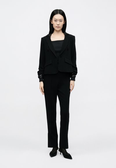 Fekete öltöny, rövidített kabáttal, kapucnival és két első zsebbel, hozzáillő, szűk nadrággal és hegyes orrú fekete magassarkúval.