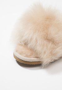 Chaussons en fausse fourrure beige avec une tige texturée, un embout en daim doux et une semelle en caoutchouc avec une base beige et des coutures blanches.