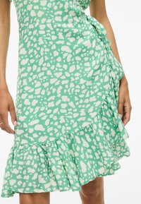 Grønn omslagskjole laget av lett materiale med et hvitt abstrakt mønster. Har en volangkant og en sidesnøring.