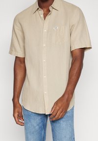 Uomo che indossa una camicia beige a maniche corte con bottone e tasca sul davanti, accompagnata da jeans azzurri, in piedi contro uno sfondo neutro.