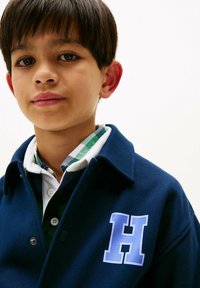 Ragazzo con capelli corti e scuri che indossa una giacca blu navy con una patch "H" blu su una camicia a quadri verde e bianca.
