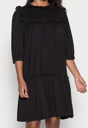 Mujer con un vestido negro suelto con mangas tres cuartos y detalles de encaje en el canesú y el cuello.