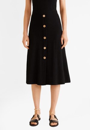 Jupe midi noire avec boutons en bois sur le devant, portée avec des sandales noires à bout ouvert et une cheville de coquillages à la cheville gauche.
