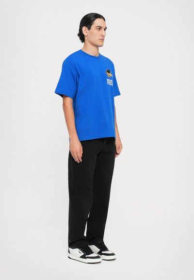 Blå bomullt-skjorte med korte ermer og rund hals, med en trykt logo på brystet. Bært med svarte bukser og svart-hvite sneakers.