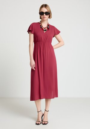 Frau in einem dunkelroten Midikleid mit flatternden Ärmeln, schwarzen Sandalen mit hohen Absätzen, massiver Halskette und schwarzen Sonnenbrillen, die vor einem schlichten Hintergrund steht.