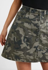 Närbild av en kjol med kamouflage-mönster, knäppt framtill, som bärs av en person med en svart topp. Kjolen har framfickor och bältesöglor.