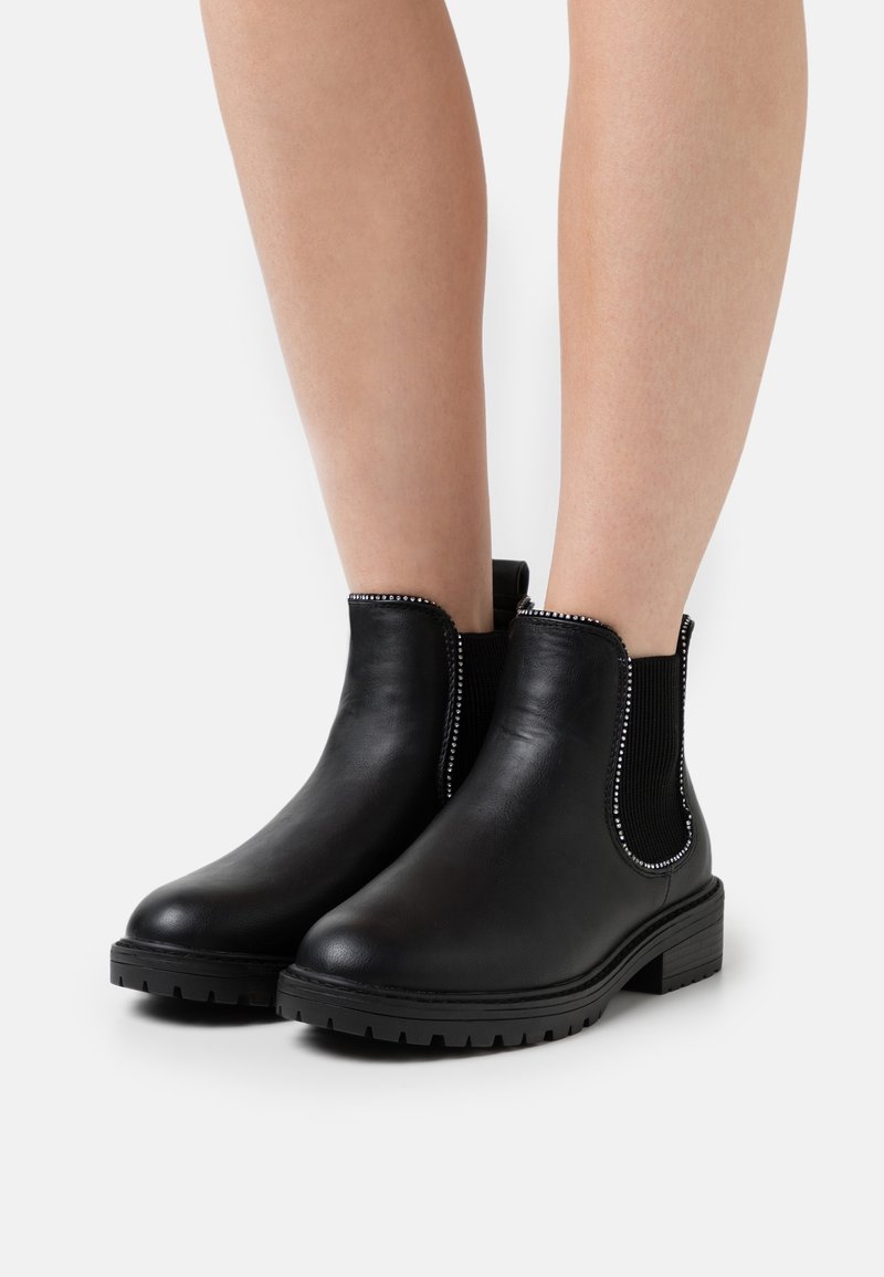 Bottines en cuir noir avec des strass décoratifs le long des panneaux latéraux élastiques, portées sur jambes nues sur fond blanc.