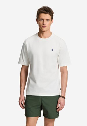 Jonge man die een effen wit T-shirt met korte mouwen en een klein donker logo draagt en donkergroene shorts tegen een effen lichte achtergrond.