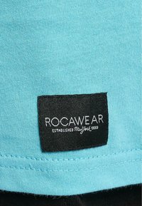 Aqua spalvotas medvilnės audinys su briaunotu paviršiumi, būdingas juodas etiketė su balta užrašu "ROCAWEAR" ir "ĮSTEIGTA Niujorke 1999".