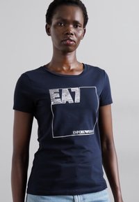 Tumedast puuvillane T-särk, millel on valge graafiline disain koos "EA7" tekstiga ja "EMPORIO ARMANI" logoga allosas. Klassikaline ümmargune kaelus.