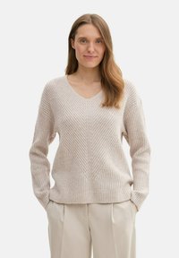 Ljus beige stickad tröja med V-ringning, med ett texturerat mönster och långa ärmar. Tillsammans med ljusa byxor.