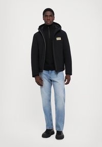 Jachetă neagră cu glugă și fermoar frontal, cu o pată de logo aurie, purtată peste un pulover negru cu guler înalt. Jeans albastri deschiși și încălțăminte neagră completează ținuta.