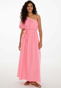 Maxi šaty v jasne ružovej látke, s jedným vypnutým ramienkom a balónovou rukávom, so šnúrkou v páse a splývavým strihom až po členky.