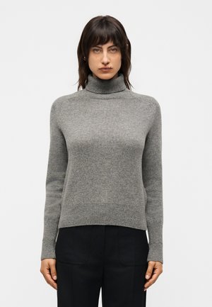 Žena u sivoj rolki i crnim hlačama stoji na svijetloj jednobojnoj pozadini, okrenuta prema naprijed.