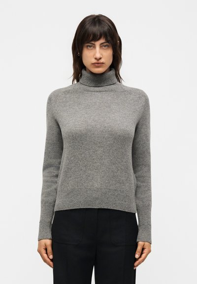 Femme portant un pull à col roulé gris et un pantalon noir, debout devant un fond clair uni, face à l’avant.