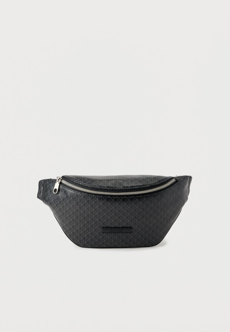 Schwarze Calvin Klein Bauchtasche mit dezentem, sich wiederholendem Logomuster und silbernem Reißverschluss auf weißem Hintergrund.