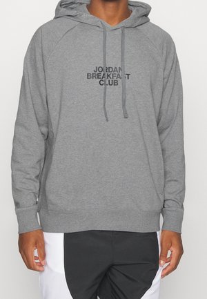 Person iført en grå hættetrøje med "JORDAN BREAKFAST CLUB" trykt sort på brystet og sort-hvide bukser.