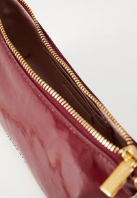 Burgundy bőrzacskó fényes felülettel, cipzáras tetejével, aranyszínű fém részletekkel és diszkrét belső zsebbel a rendezettség érdekében.