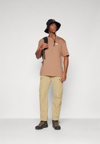 T-shirt en coton marron avec logo, pantalon beige avec poches zippées, chapeau seau noir et chaussures de randonnée noires avec accents jaunes.