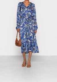 Frau trägt ein blaues, florales Mittelkleid mit langen Ärmeln und einem Taillengürtel, hält eine braune Lederhandtasche und trägt braune Sandalen mit Riemchen.
