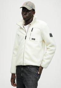 Veste en polaire blanche avec fermeture éclair et surface texturée, dotée d'une poche poitrine, d'accents de fermeture éclair noirs et d'un patch sur la manche.