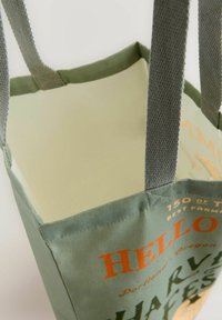 Borsa tote riutilizzabile in tela verde con manici in rete. L'interno è di colore chiaro, con testo arancione e verde scuro e un design a tema agricolo.