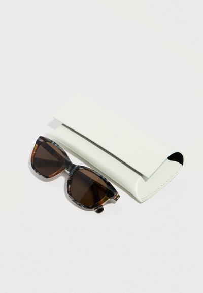 Lunettes de soleil à motif écaille avec des verres foncés placées à côté d'un étui rigide blanc fermé sur un fond uni.