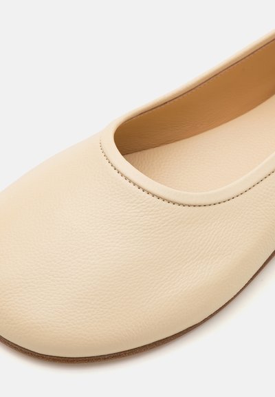 Ballerine en cuir beige avec un bout rond, un design épuré, une texture subtile et des détails de couture minimalistes près de l'ouverture à la cheville.